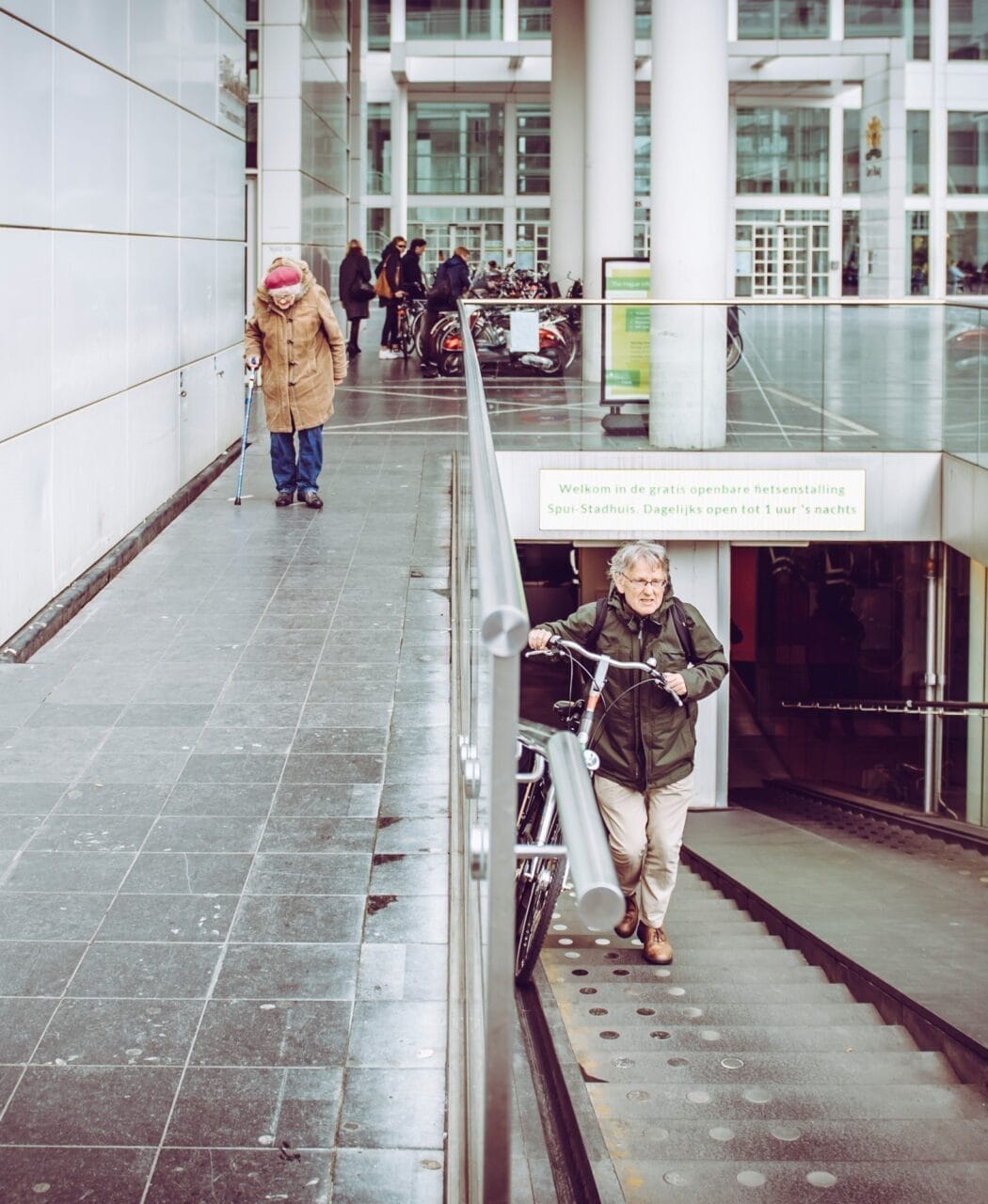 Elderly woman walking with a cane and older man walking up stairs with a bicycle in a building. The Hague, Netherlands